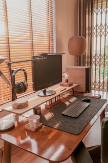 A neatly organized home office setup with a modern aesthetic. The desk features a computer monitor mounted on a raised stand, a keyboard and mouse placed on a felt mat, and a pair of black headphones hanging from a side mount. There is a cup on a coaster, a small lamp, and a decorative wooden object on the desk. Natural light filters through the wooden blinds, illuminating the space with a warm glow.