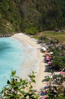 A picturesque beach with white sand and turquoise water is bordered by lush green vegetation and palm trees. Colorful umbrellas and lounge chairs are arranged along the shoreline. A few people can be seen enjoying the water and relaxing on the beach.