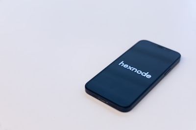 A smartphone is placed on a light-colored surface with the screen displaying the logo 'hexnode'. The phone is positioned diagonally with its top left corner facing upward.