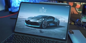 A tablet with a keyboard is displaying a car wallpaper showing a sleek, futuristic sports car parked on a barren landscape. The car features sharp lines and a glossy finish. On the screen, there is also a digital clock and some text in a non-English script. Surrounding the tablet are a smartphone, a pair of headphones, and a blurred background with some abstract shapes.