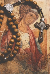 A religious painting depicts a figure with curly hair in a traditional pose. A rosary with wooden beads and an ornate cross draped over the image adds depth and texture.