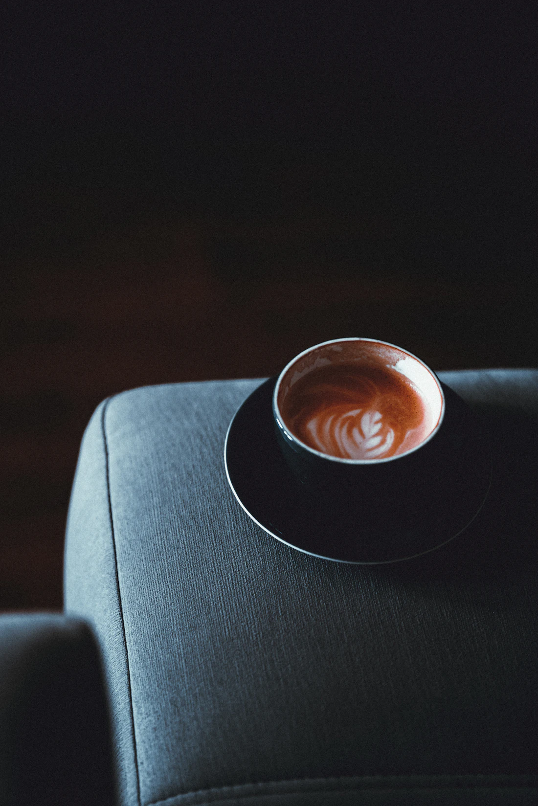 Xícara de café apoiada no braço de um sofá em tecido, em ambiente de luz baixa