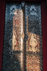 Intricately painted wooden doors featuring ornamental and symmetrical designs. The artwork displays figures with traditional attire in a symmetrical pattern, surrounded by vine-like motifs. The overall design includes ornate carvings with reflective or metallic paint creating a shimmering effect.