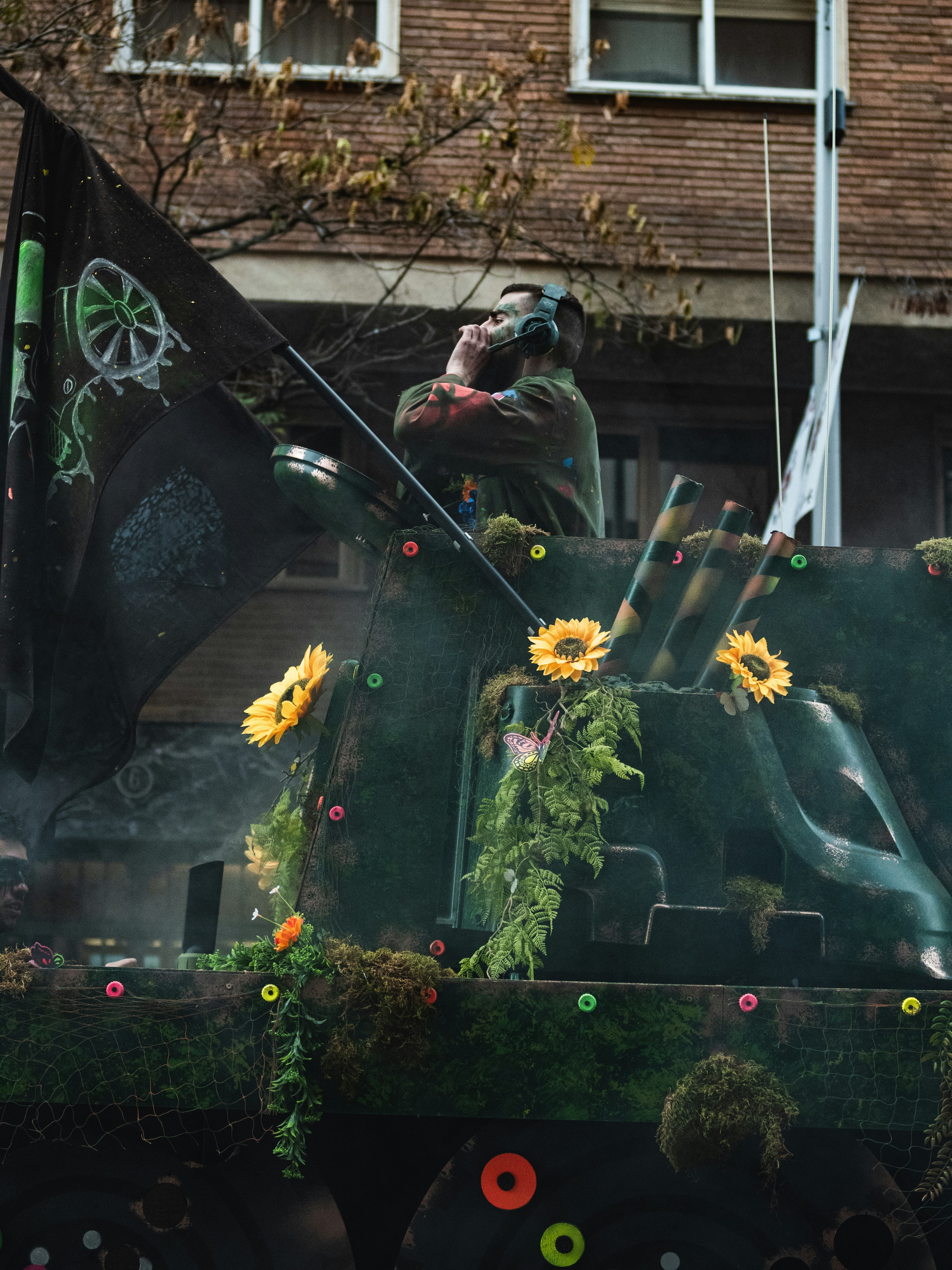 Parade float decorated with sunflowers and ivy carries a performer wearing headphones against a brick backdrop.