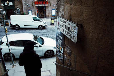 A cobblestone street scene featuring a white van and a white car parked near a stone building with a blue and red sign for ‘Bertie's Proper Fish & Chips’. A person dressed in black stands by the entrance to a narrow staircase leading down. Nearby, there is a sign indicating directions to Castlehill and The Castle, and another sign with restaurant menus.