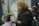A barber with curly hair and a beard is cutting the hair of a seated man with a gray man bun. Another barber is visible in the background working on a different client. The atmosphere appears to be relaxed and professional.