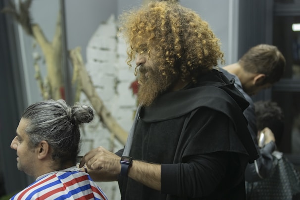 A barber with curly hair and a beard is cutting the hair of a seated man with a gray man bun. Another barber is visible in the background working on a different client. The atmosphere appears to be relaxed and professional.