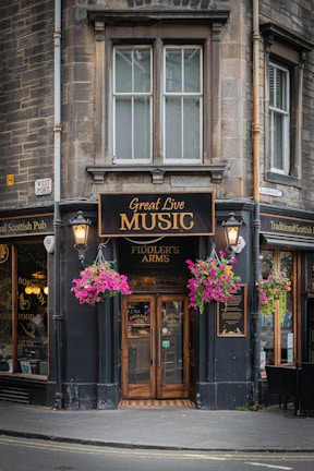 A quaint corner pub with stone exterior, featuring signage promoting live music. The building has large windows and wooden double doors adorned with the name 'Fiddler's Arms'. Hanging baskets filled with vibrant pink and purple flowers flank the entrance, and traditional street lamps are affixed near the doorway. Adjacent windows display various advertisements and decorations.