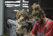 Two barbers are focused on cutting hair in a salon setting. Both individuals are attending to different clients, using scissors and other tools. The ambient lighting gives a calm and concentrated atmosphere. The background includes reflections from mirrors and other salon equipment.