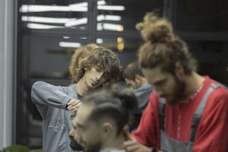 Two barbers are focused on cutting hair in a salon setting. Both individuals are attending to different clients, using scissors and other tools. The ambient lighting gives a calm and concentrated atmosphere. The background includes reflections from mirrors and other salon equipment.
