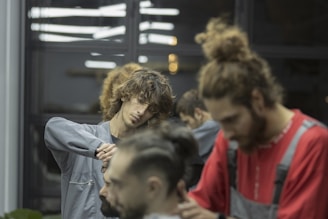 Two barbers are focused on cutting hair in a salon setting. Both individuals are attending to different clients, using scissors and other tools. The ambient lighting gives a calm and concentrated atmosphere. The background includes reflections from mirrors and other salon equipment.