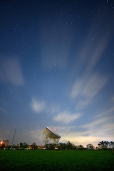 A large radio telescope dish stands prominently against a starry night sky, with blurred clouds moving swiftly across the horizon. The dish is surrounded by a grassy field and silhouetted trees, while city lights create a faint glow near the horizon.