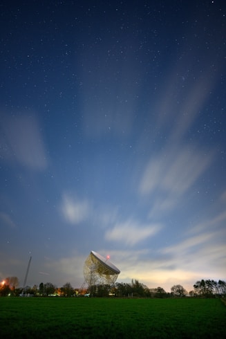 A large radio telescope dish stands prominently against a starry night sky, with blurred clouds moving swiftly across the horizon. The dish is surrounded by a grassy field and silhouetted trees, while city lights create a faint glow near the horizon.