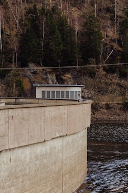 A concrete dam structure curves over a body of water with trees growing densely on the rocky hillside in the background. A small building with a flat roof and grid windows is situated on top of the dam. Two people are leaning over the railing, gazing at the water below.