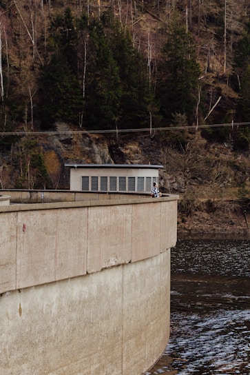 A concrete dam structure curves over a body of water with trees growing densely on the rocky hillside in the background. A small building with a flat roof and grid windows is situated on top of the dam. Two people are leaning over the railing, gazing at the water below.