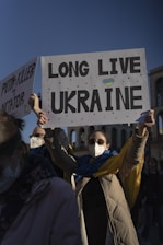 A person wearing a mask, sunglasses, and a long coat is holding a sign that says 'LONG LIVE UKRAINE' with a small heart featuring the colors of the Ukrainian flag. Another sign partially visible reads 'PUTIN = KILLER AND A DICTATOR.' The background features ancient architectural structures under a clear blue sky.