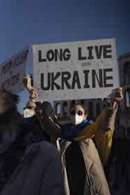 A person wearing a mask, sunglasses, and a long coat is holding a sign that says 'LONG LIVE UKRAINE' with a small heart featuring the colors of the Ukrainian flag. Another sign partially visible reads 'PUTIN = KILLER AND A DICTATOR.' The background features ancient architectural structures under a clear blue sky.