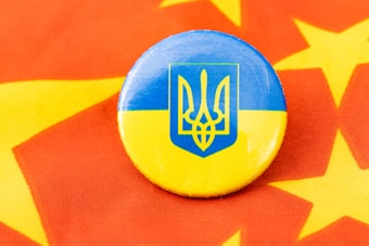 A round badge featuring the blue and yellow flag of Ukraine with the national coat of arms is placed on a red fabric decorated with yellow stars.