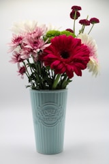 A bouquet of mixed flowers including red, white, pink, and green blooms is arranged in a tall, pastel blue vase with a decorative embossed design that reads 'FLOWERS & GARDEN'. The vase is placed against a simple, light background, emphasizing the vibrant colors of the flowers.
