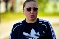 A person with sunglasses is wearing a dark tracksuit top with white stripes and a logo on the front. The background is blurred, with shades of green indicating an outdoor setting.