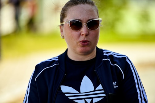 A person with sunglasses is wearing a dark tracksuit top with white stripes and a logo on the front. The background is blurred, with shades of green indicating an outdoor setting.