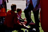 A person is sitting on the ground, possibly after a sporting event, and is being attended to by another individual who appears to be providing assistance. There are several other people nearby, one of whom is partially visible. The setting is an outdoor area with grass and pavement.