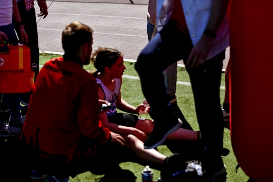A person is sitting on the ground, possibly after a sporting event, and is being attended to by another individual who appears to be providing assistance. There are several other people nearby, one of whom is partially visible. The setting is an outdoor area with grass and pavement.