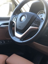 A close-up of a car's steering wheel featuring the BMW logo prominently in the center, surrounded by mounted control buttons. The interior displays leather upholstery with a mix of black and brown colors, hinting at a luxury car design. The blurred image in the background suggests the dashboard, possibly with a speedometer visible.