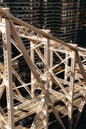 An intricate metal bridge structure set against a backdrop of tall, dark residential buildings. The bridge features crisscrossing beams and trusses with a light, aged patina. Below, vehicles are visible driving along the road that runs parallel to the bridge.