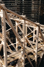 An intricate metal bridge structure set against a backdrop of tall, dark residential buildings. The bridge features crisscrossing beams and trusses with a light, aged patina. Below, vehicles are visible driving along the road that runs parallel to the bridge.