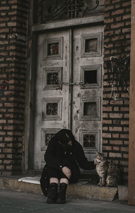 A person dressed in dark clothing sits on a stoop in front of an old, weathered door made from wood and surrounded by a brick wall. The individual is gently petting a tabby cat that stands next to them. The scene conveys a sense of serenity and companionship.