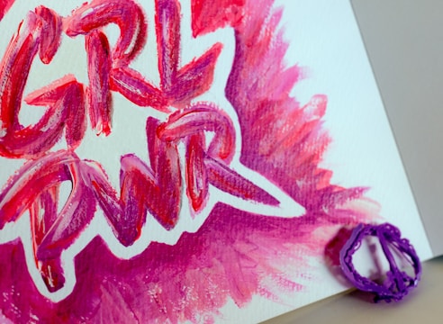 Painted words 'GRL PWR' in bold, vibrant strokes of pink, purple, and red on a white canvas. A purple plastic object is placed near the edge of the canvas.