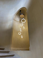 A minimalist and elegant lighting fixture consisting of multiple suspended circular lights hangs in a stairwell with smooth, curved walls. The warm tones of the walls complement the modern design of the fixture, creating a serene and sophisticated atmosphere.