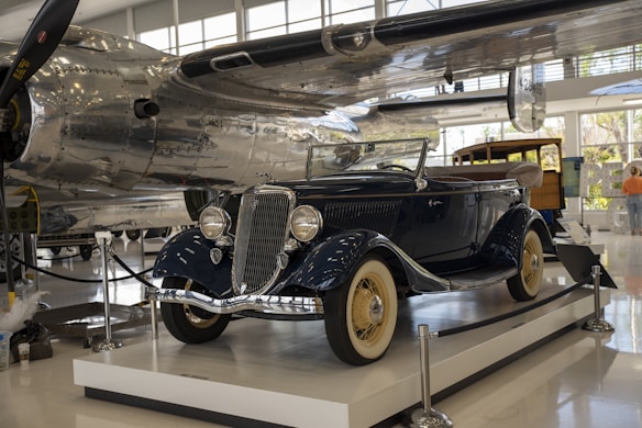 A vintage car with a polished navy blue exterior and beige wheels is displayed in an indoor museum. The vehicle is roped off with black stanchions and is positioned on a white platform. In the background, parts of an airplane with shiny metallic surfaces are visible, along with some museum displays and signage.