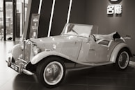 An elegant vintage car is displayed indoors, featuring a classic design with chrome accents and distinctive curves. The convertible has a prominent front grille and spoked wheels. The background includes modern architecture and a sign with Chinese characters.
