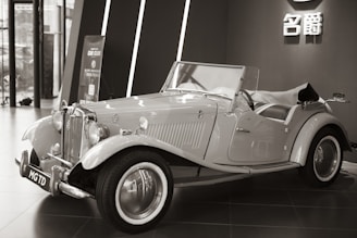 An elegant vintage car is displayed indoors, featuring a classic design with chrome accents and distinctive curves. The convertible has a prominent front grille and spoked wheels. The background includes modern architecture and a sign with Chinese characters.