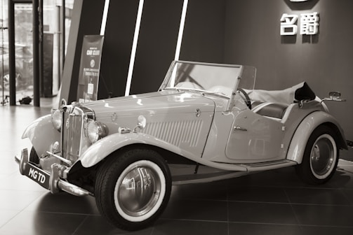 An elegant vintage car is displayed indoors, featuring a classic design with chrome accents and distinctive curves. The convertible has a prominent front grille and spoked wheels. The background includes modern architecture and a sign with Chinese characters.