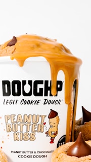 A container labeled 'DOUGHP' featuring 'Peanut Butter Kiss' cookie dough with peanut butter and chocolate flavor. Peanut butter drips down the side of the container, and cookies with chocolate kisses are visible.