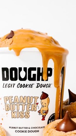 A container labeled 'DOUGHP' featuring 'Peanut Butter Kiss' cookie dough with peanut butter and chocolate flavor. Peanut butter drips down the side of the container, and cookies with chocolate kisses are visible.