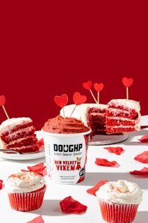 A vibrant scene featuring a red velvet themed dessert spread. The centerpiece is a container of 'Doughp Red Velvet Vixen' cookie dough surrounded by slices of red velvet cake adorned with cream frosting and heart-shaped decorations. Two red velvet cupcakes with white frosting are placed in the foreground, along with scattered red rose petals.