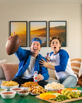 Two young men sitting on a sofa look excited as they engage in watching something, possibly a sports game. One of them, wearing a blue cap and jacket, holds an American football and a can of soda. The other, wearing a blue and white baseball shirt, appears surprised and is also holding a can. In front of them, there is a table covered with various snacks, including chips, salsa, pretzels, popcorn, and a vegetable platter. Three framed photographs of a landscape hang on the wall in the background.