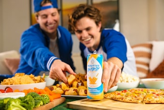 Two people are smiling and reaching for snacks on a table filled with various foods, including pizza, popcorn, chips, fresh vegetables, and a can of energy drink labeled 'Positive Energy'. The setting appears relaxed and casual.