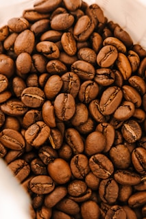 A close-up view of a pile of roasted coffee beans, showcasing their rich brown color and textured surface. The beans are tightly packed together, with visible creases and natural imperfections, emphasizing their freshness and quality.