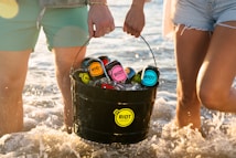 Two people are carrying a black bucket filled with colorful cans of Riot Energy drinks, with ice keeping them cool. They are standing in shallow ocean water, and the sunlight creates a warm and refreshing summer setting.
