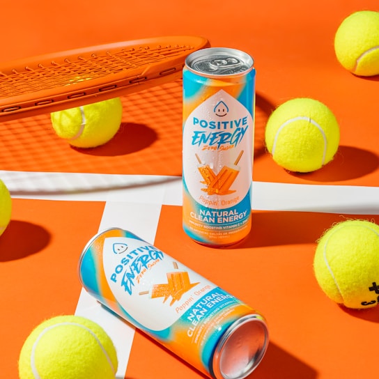 Two cans of Positive Energy drink in a vibrant setting, surrounded by several yellow tennis balls on an orange and white background. An orange tennis racket is positioned to the side, creating a sporty atmosphere.