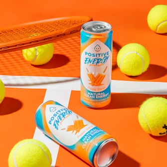Two cans of Positive Energy drink in a vibrant setting, surrounded by several yellow tennis balls on an orange and white background. An orange tennis racket is positioned to the side, creating a sporty atmosphere.