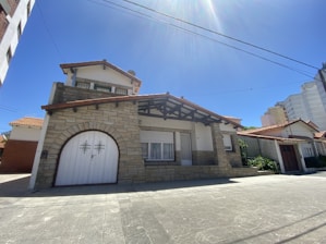 A two-story house with stone facade, a large white wooden garage door, and a prominent gabled roof. The building features a mix of red brick and white plaster accents, with shuttered windows. Surrounding the house are other residential structures, and the sky is clear blue and sunny.