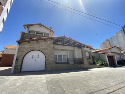 A two-story house with stone facade, a large white wooden garage door, and a prominent gabled roof. The building features a mix of red brick and white plaster accents, with shuttered windows. Surrounding the house are other residential structures, and the sky is clear blue and sunny.