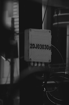 A black and white image of an electrical junction box mounted on a vertical pole. The box is labeled with the code '2DJ030305' and has several wires and cables connected beneath it. The background is blurred, with indistinct structures visible.