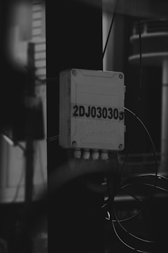 A black and white image of an electrical junction box mounted on a vertical pole. The box is labeled with the code '2DJ030305' and has several wires and cables connected beneath it. The background is blurred, with indistinct structures visible.